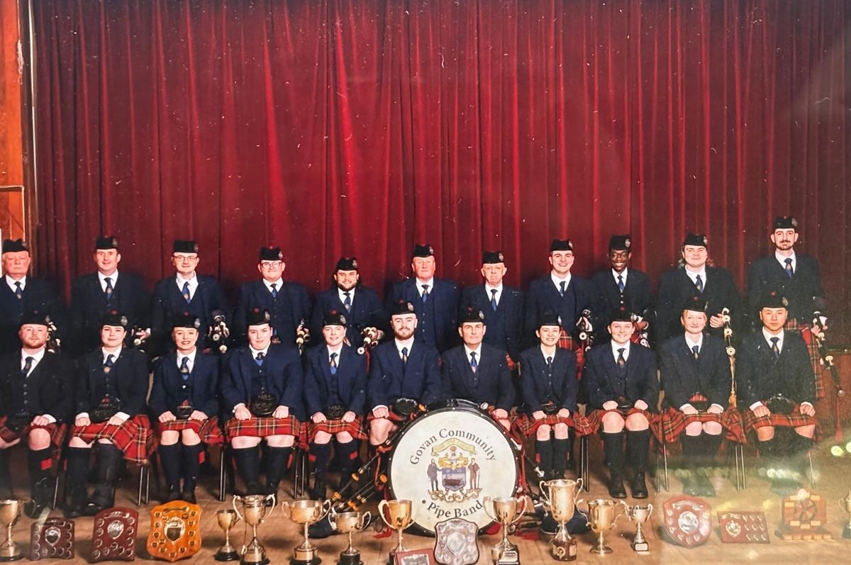 The band seated in formal uniform in front of a red curtain with their trophies laid out at their feet